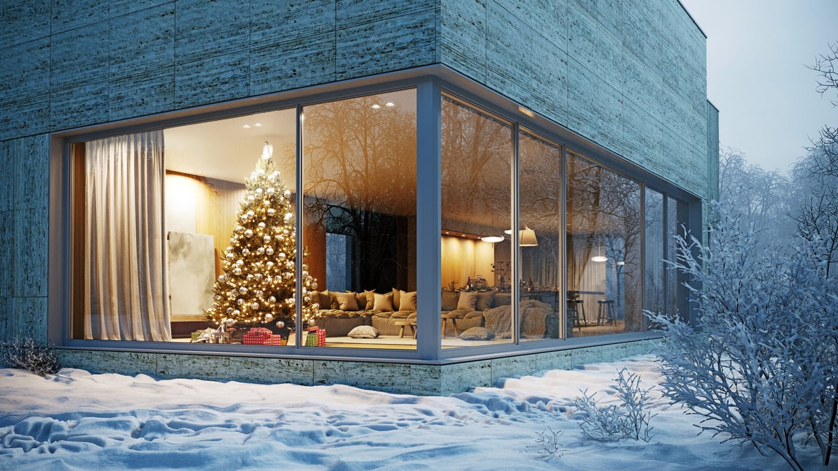 A modern house with large glass windows reveals a cozy living room with a lit Christmas tree at twilight