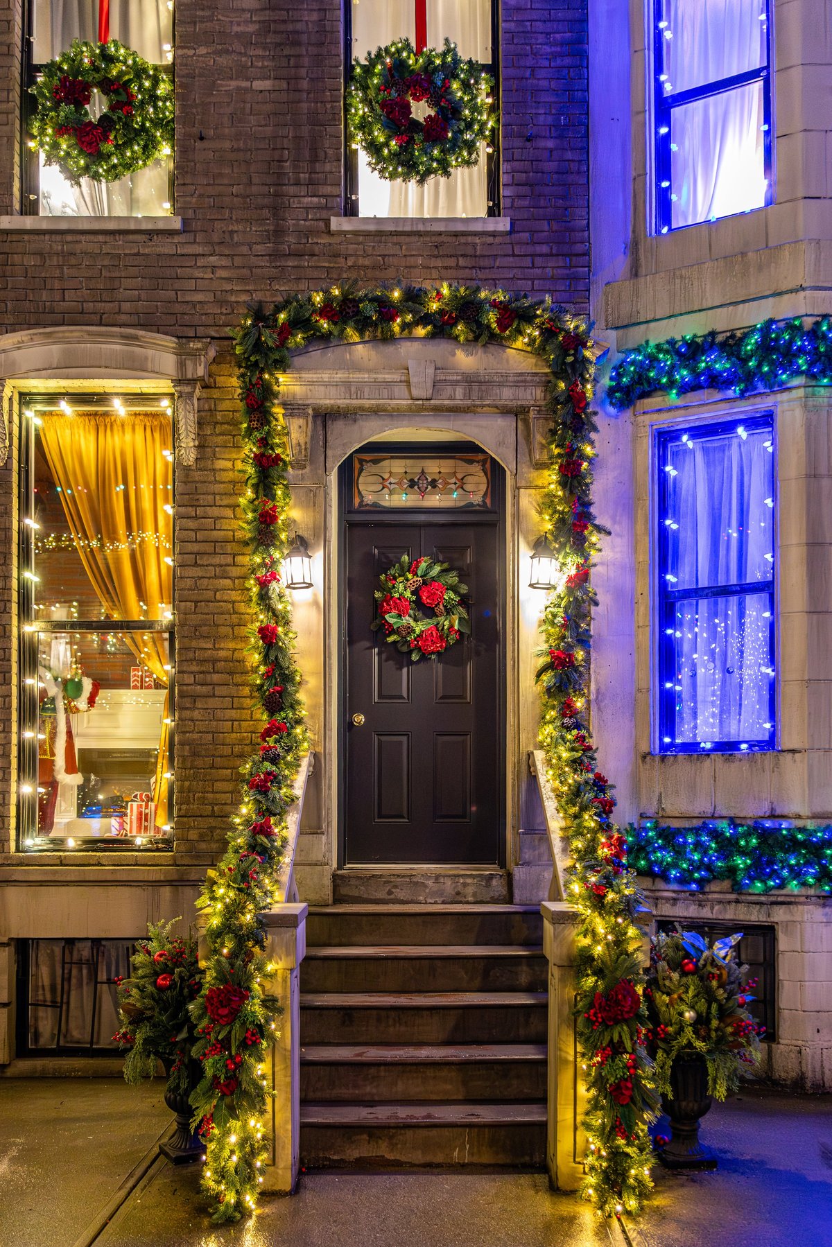 Beautifully decorated brick townhouse entrance at night featuring Christmas wreaths, evergreen garlands, and warm glowing holiday lights