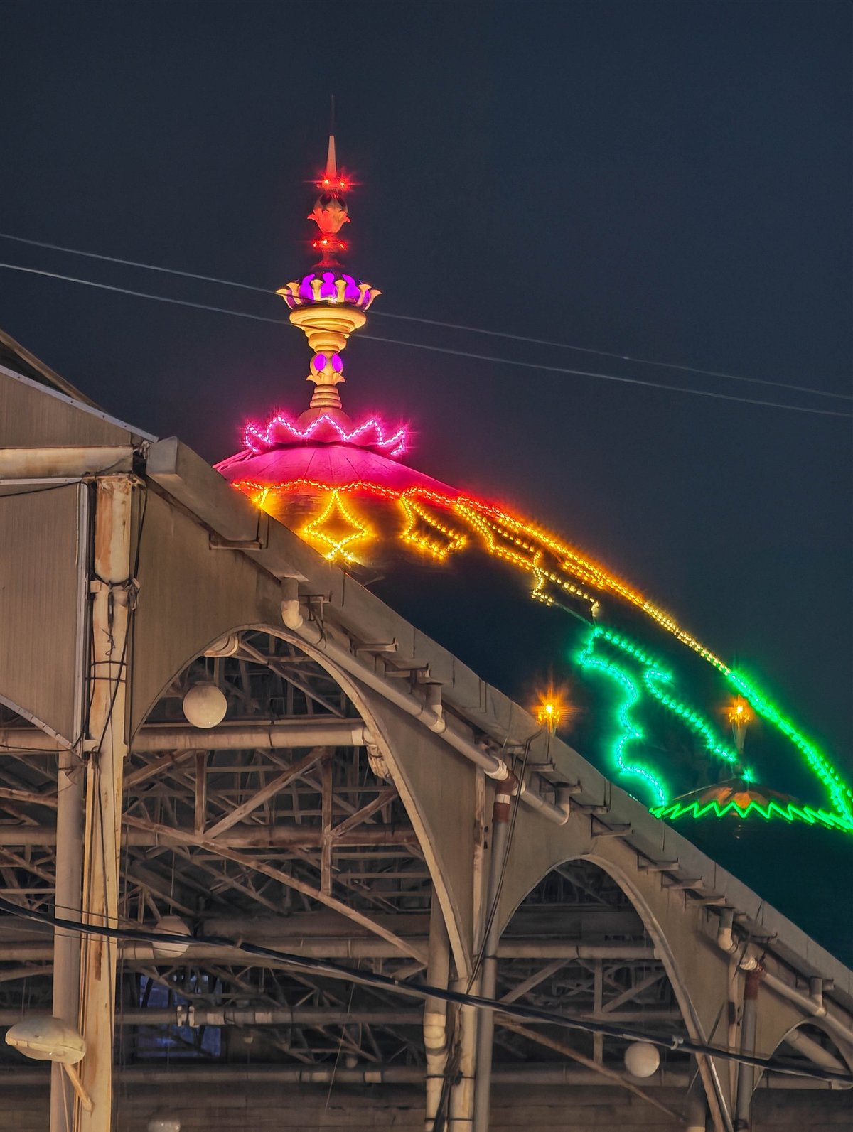 Ornate illuminated spire and colorful light arches above structure at night