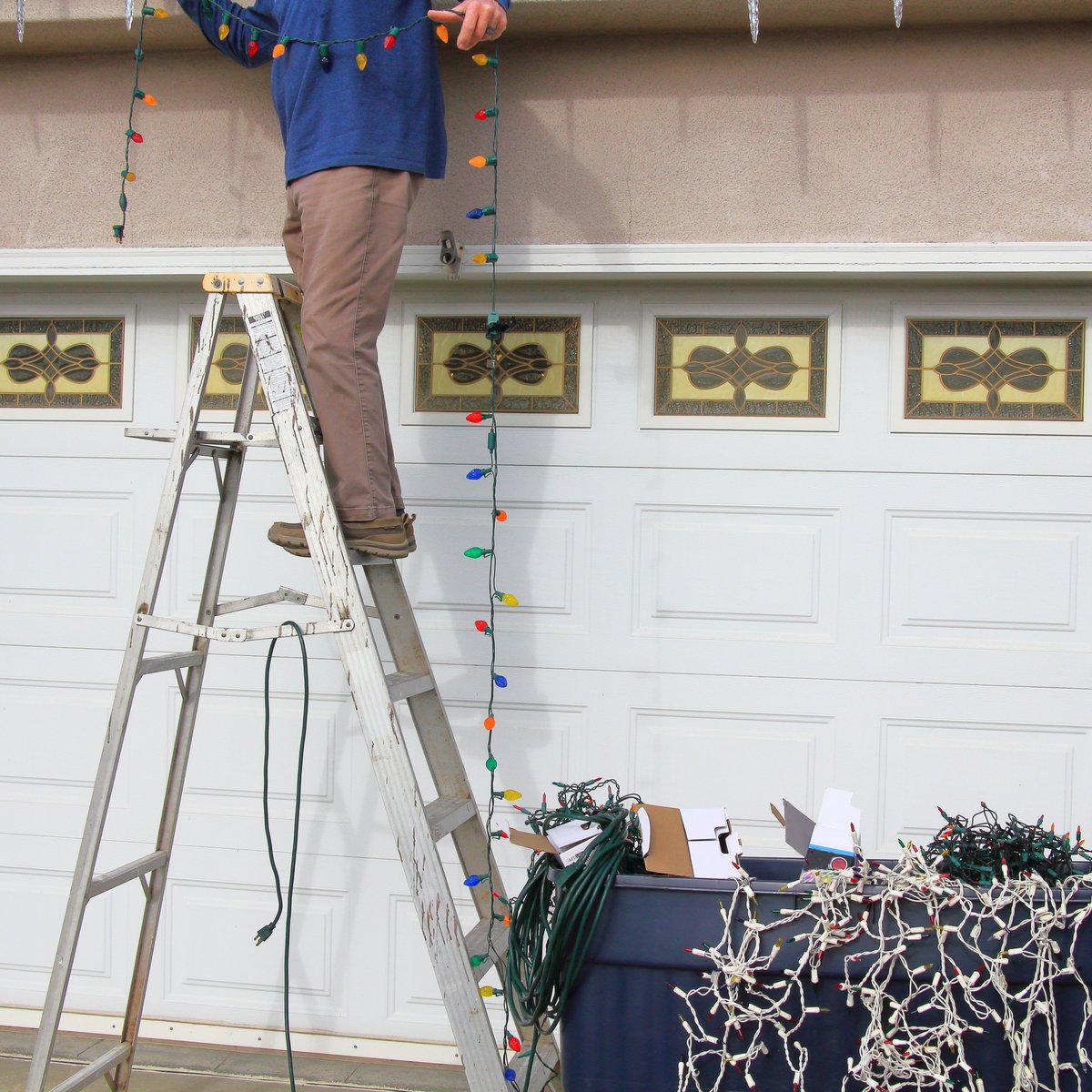 Worker balancing on ladder stretching Christmas lights along the side of a house